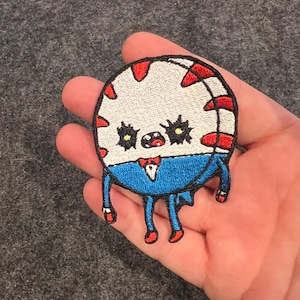 May include: A round, embroidered patch featuring a cartoon character with a white head, red accents, and a blue suit. The character has black eyes, a red bow tie, and red shoes. The patch is held in a hand.