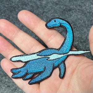 Peut inclure: Un écusson brodé représentant un plésiosaure bleu avec un long cou et des nageoires. La créature est représentée avec un motif de vague bleu clair horizontal sur le corps et est soulignée par un fil noir sur un fond texturé.