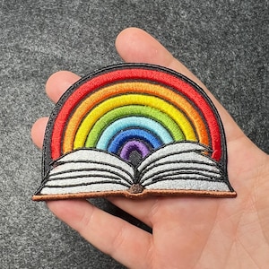 May include: A rainbow-colored embroidered patch shaped like an open book. The rainbow is made of red, orange, yellow, green, blue, indigo, and violet. The book is open and has white pages with black lines.