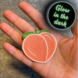 May include: An iron-on patch embroidered with a peach in a peach colour, with a green stem and leaf. The patch glows in the dark. "Glow in the dark" is written in green text on a black circle.