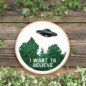 Könnte beinhalten: Ein weißer Stickrahmen mit einem Kreuzstich-Design eines UFOs, das über einen Wald fliegt, mit dem Text "I WANT TO BELIEVE".