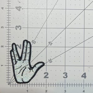 May include: A white and silver embroidered patch of a hand making the Vulcan salute, a popular gesture from the Star Trek franchise.