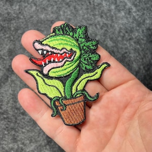 May include: Embroidered patch featuring a green, cartoon-style plant with a large, open mouth, red tongue, and white teeth. The plant is in a brown pot with green leaves and a green stem. The patch is designed with black outlines.