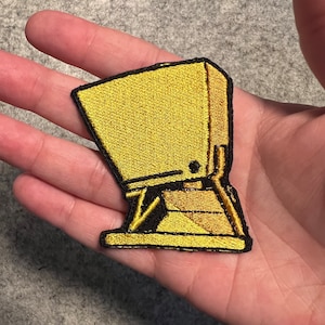 May include: A gold embroidered patch of a cartoon character with a box-like head and legs. The character is standing on a platform.