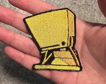 May include: A gold embroidered patch of a cartoon character with a box-like head and legs. The character is standing on a platform.