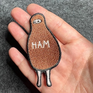 May include: An embroidered patch shaped like a brown ham with a black border. The patch features a small white face at the top, the word HAM stitched in white thread in the center, and two small white legs extending from the bottom of the figure.