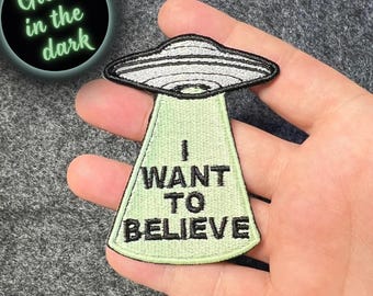 May include: Embroidered patch featuring a UFO design with the text "I WANT TO BELIEVE" in black. The UFO is gray and black, with a green beam. The patch glows in the dark.
