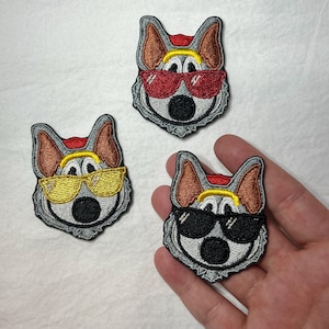 May include: Three embroidered iron-on patches featuring a cartoon dog wearing sunglasses. The dog has a gray face, brown ears, and a red bandana. The sunglasses are yellow, red, and black.