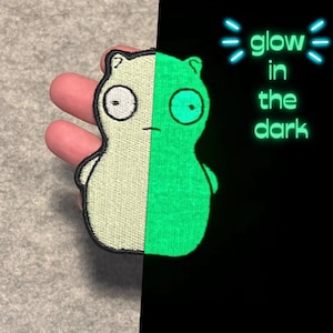 Pode incluir: Um remendo bordado branco e verde de um personagem de desenho animado. O personagem é de cor verde claro quando exposto a uma luz negra. O texto "glow in the dark" está escrito em branco sobre um fundo preto.