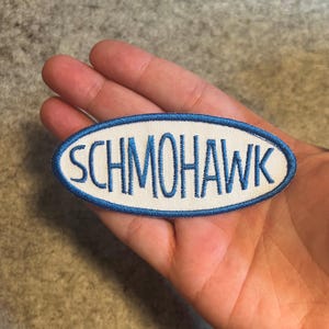 May include: A white oval patch with a blue border. The text "SCHMOHAWK" is embroidered in blue on the patch.