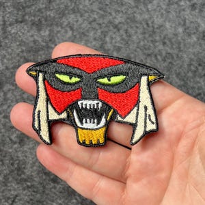 Peut inclure: Écusson brodé représentant un visage stylisé avec un masque rouge, une coiffe noire et des yeux jaunes. Le patch a une bordure blanche à beige et une bouche jaune et blanche avec des dents pointues. Pour vêtements ou accessoires.
