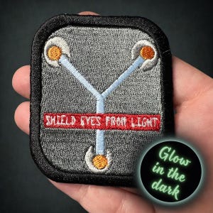 May include: Embroidered patch with a gray background, black border, and the text "SHIELD EYES FROM LIGHT" in red. Features a blue Y-shaped design with orange and white accents. A circular "Glow in the dark" graphic is in the lower right.