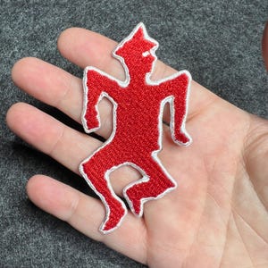 Op de afbeelding: Een rode geborduurde patch in de vorm van een gestileerde figuur met een witte omtrek. De figuur staat in een rennende pose, met uitgestrekte armen en gebogen benen. De patch wordt in een hand gehouden.