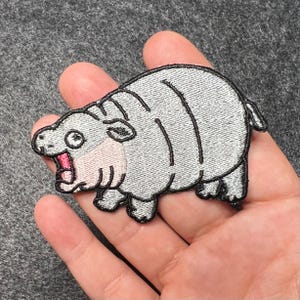 May include: A gray embroidered iron-on patch of a cartoon hippopotamus with an open mouth and pink inside. The hippopotamus has black eyes and a black outline.