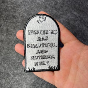 Pode incluir: Um remendo bordado em prata e preto em forma de lápide com o texto "EVERYTHING WAS BEAUTIFUL AND NOTHING HURT".