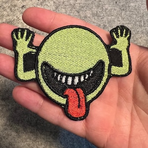 May include: A green and black embroidered patch of a cartoon face with a wide, toothy grin and a red tongue sticking out. The face has two arms raised in the air.