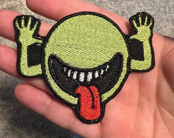 May include: A green and black embroidered patch of a cartoon face with a wide, toothy grin and a red tongue sticking out. The face has two arms raised in the air.
