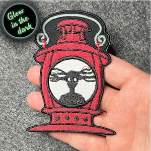 May include: Red and black embroidered iron-on patch of a lantern with a creature inside. The lantern has a handle and a round glass window. The creature is black with long, thin arms and a white face. The text "Glow in the dark" is visible in the upper left corner of the image.
