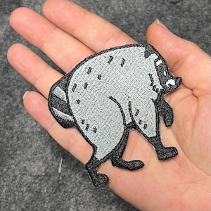 以下が含まれることがあります： アライグマの形をした刺繍ワッペン。ライトグレーの体に、顔、足、縞模様の尻尾が黒で縁取られています。背中には小さな黒いダッシュ状の模様があり、鼻先には白い斑点があしらわれたデザインです。