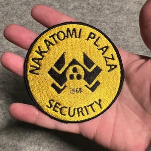 Puede incluir: Un parche redondo bordado en oro con texto negro que dice "Nakatomi Plaza Security". El parche presenta un logotipo negro estilizado de un edificio con una cerradura en el centro.