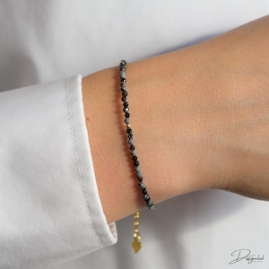 Könnte beinhalten: Ein zartes Perlenarmband mit kleinen schwarzen und grauen Perlen. Das Armband hat einen goldfarbenen Verschluss und einen kleinen Anhänger. Das Armband wird am Handgelenk getragen, mit einem weißen Hemd im Hintergrund.
