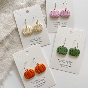 May include: Four pairs of pumpkin-shaped earrings in cream, pink, orange, and green. Each pair is on a white card with silver hoops and a gold-coloured stem. The cards read "CRAFTED BY JJ CO." and include a website address.