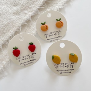 Peut inclure: Trois paires de boucles d'oreilles en forme de fruits : fraises, pêches et citrons. Les boucles d'oreilles sont sur des cartes blanches avec le texte "CRAFTED BY JJ. CO. Handmade with joy" et des réseaux sociaux.