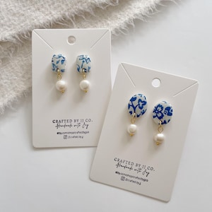 Può includere: Due paia di orecchini su cartoncini bianchi. Gli orecchini presentano un motivo floreale blu e bianco con una goccia di perla bianca. Le carte riportano il testo "CRAFTED BY JJ. CO. Handmade with Joy" e informazioni sui social media.