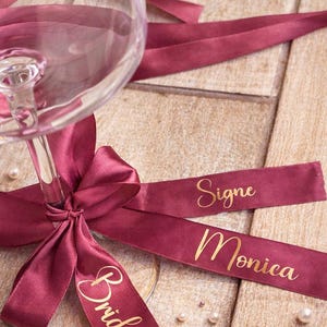 May include: A wine glass with a claret ribbon tied in a bow. The ribbon has the word "Bride" printed on it. Two additional ribbons are visible, one with the name "Signe" and the other with the name "Monica". Small pearls are scattered around the glass.
