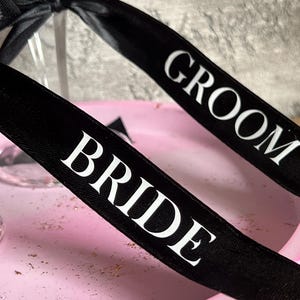May include: Two black satin ribbons tied in bows around clear glass stems. One ribbon features the white text GROOM and the other features the white text BRIDE. The ribbons are positioned over a light pink, textured, circular base.