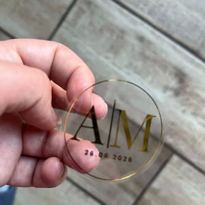 Könnte beinhalten: Transparenter, runder Aufkleber mit goldfarbenem Schriftzug und goldenem Rand. Die Initialen "AM" sind vertikal dargestellt, darunter das Datum "26.06.2026". Der Aufkleber wird in einer Hand gehalten.