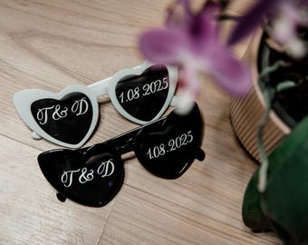 May include: Two pairs of heart-shaped sunglasses, one white and one black. The white sunglasses have "T & D" and "1.08.2025" written on them. The black sunglasses have "T & D" and "1.08.2025" written on them.