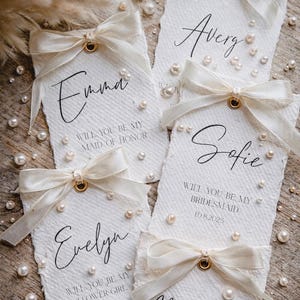 Puede incluir: Etiquetas de papel blanco texturizado con nombres en escritura elegante, adornadas con lazos de color marfil y perlas de imitación. Las etiquetas dicen "¿Quieres ser mi dama de honor?", "Niña de las flores" y "Dama de honor". Un tema de invitación de boda.