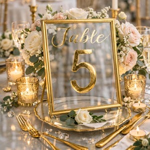 May include: A gold framed table number 5, with the word "Table" above the number. The table setting includes gold cutlery, candles, and floral arrangements with white and pink roses. The table is set for a special event.