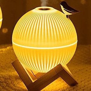 May include: A white ceramic diffuser with a wooden base and a bird-shaped handle. The diffuser is emitting a white mist and has a warm, yellow glow.