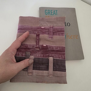 Può includere: Una copertina di libro patchwork nei toni del marrone e del bordeaux, che ricorda i jeans. La copertina ha passanti per cintura e un aspetto strutturato. Sullo sfondo è visibile un libro con la parola "GREAT".