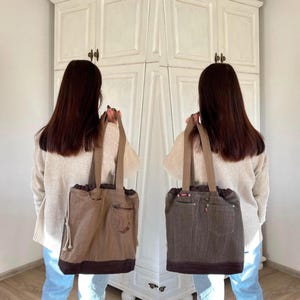 Op de afbeelding: Twee bruine tote tassen met denim zakken en bruine suède-achtige rand. De tassen hebben beige bandjes en een trekkoordsluiting. De tassen worden voor een witte kast gehouden.