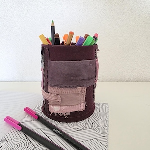 May include: A cylindrical, burgundy-colored pen holder made from repurposed denim, filled with various colored pens and pencils. Two pink-tipped pens rest on a patterned notebook. The pen holder has a patchwork design with visible stitching and a zipper.