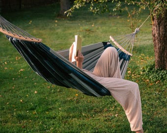 May include: A person relaxing in a hammock outdoors, reading a book. The hammock is blue and gray, suspended between trees in a grassy area. The person is wearing light-colored pants. The scene is bathed in natural light.