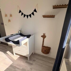 K&ouml;nnte beinhalten: Ein wei&szlig;er Wickeltisch aus Holz mit einer schwarz-wei&szlig; gemusterten Wickelauflage, einer schwarzen Stoffwimpelkette, zwei kleinen Wandregalen aus Holz, drei kleinen Elefanten-Wandhaken aus Holz und einem geflochtenen Wandkorb in Giraffenform.