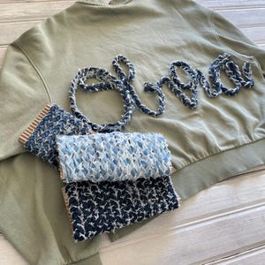 Op de afbeelding: Een olijfgroene sweatshirt met het woord "Shore" geborduurd in een denim-achtige blauwe draad. Drie stofstalen in blauwe en witte tinten worden onder het sweatshirt weergegeven.