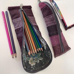 May include: Two open purple pencil cases with a floral interior, filled with colored pencils. The cases are made of patchwork fabric in shades of purple. Two pencils are next to the open case. A notebook is in the background.
