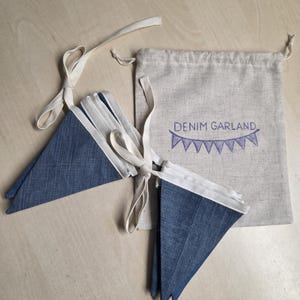 K&ouml;nnte beinhalten: Eine Denim-Wimpelkette mit dreieckigen blauen Stoffflaggen an einem wei&szlig;en Band. Die Wimpelkette wird mit einem beigen Kordelzugbeutel geliefert, der mit dem Text DENIM GARLAND und einer kleinen, violett gestickten Wimpelgrafik versehen ist.