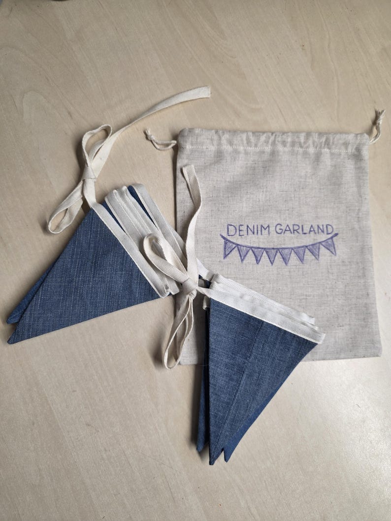 K&ouml;nnte beinhalten: Eine Denim-Wimpelkette mit dreieckigen blauen Stoffflaggen an einem wei&szlig;en Band. Die Wimpelkette wird mit einem beigen Kordelzugbeutel geliefert, der mit dem Text DENIM GARLAND und einer kleinen, violett gestickten Wimpelgrafik versehen ist.