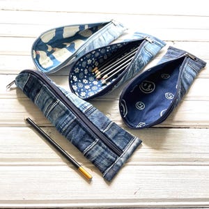 Può includere: Diversi astucci per matite in denim con diversi motivi interni sono esposti su una superficie di legno bianco. Un astuccio è aperto, rivelando matite, mentre altri mostrano disegni di balene, fiori e faccine sorridenti. Una singola matita con la punta dorata è appoggiata accanto a un astuccio chiuso.