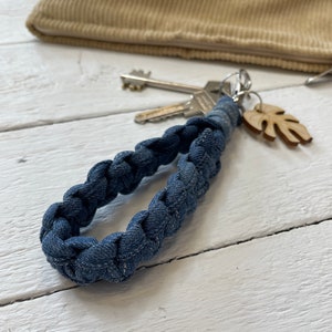 Op de afbeelding: Een blauwe denim sleutelhanger met een gevlochten ontwerp. De sleutelhanger heeft een zilveren sleutelring en een houten blaadjeshanger.