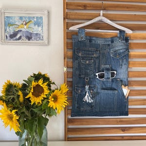 Könnte beinhalten: Ein Jeansrock mit mehreren Taschen und einem weißen Quastenanhänger an einem Schlüsselband. Der Rock hängt an einer Holzlattenwand mit einem weißen Kleiderbügel.