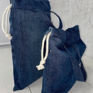 May include: Two denim drawstring bags with white drawstrings. The larger bag is lying on its side, while the smaller bag is standing upright.
