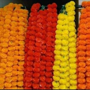May include: A close-up of five strands of marigold garlands, each strand is a different colour: orange, red, yellow, and two shades of orange. The garlands are made of artificial marigolds and are strung together with a thin string.