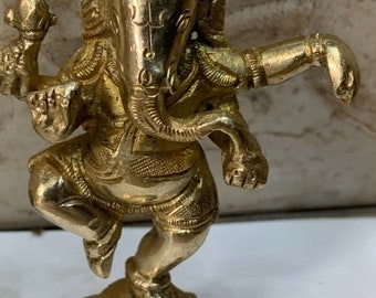 May include: A gold-colored metal statue of the Hindu deity Ganesha. The statue depicts Ganesha in a dancing pose with one hand raised and the other holding a small object. The statue is standing on a square base.